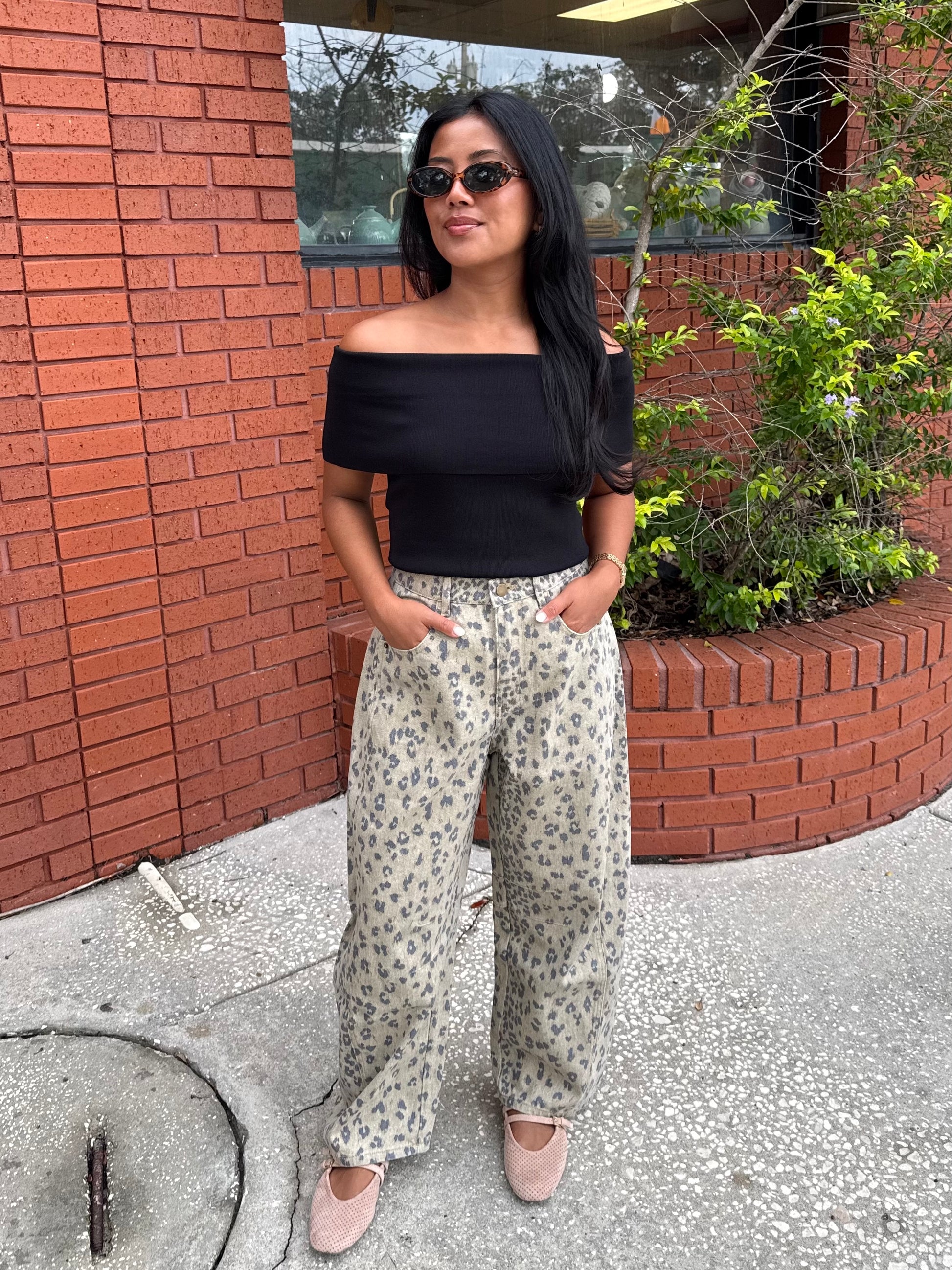 A woman wearing a black off-the-shoulder top and beige wide-leg pants with a blue floral pattern, standing outdoors in front of a red brick wall.