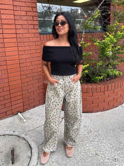 A woman wearing a black off-the-shoulder top and beige wide-leg pants with a blue floral pattern, standing outdoors in front of a red brick wall.