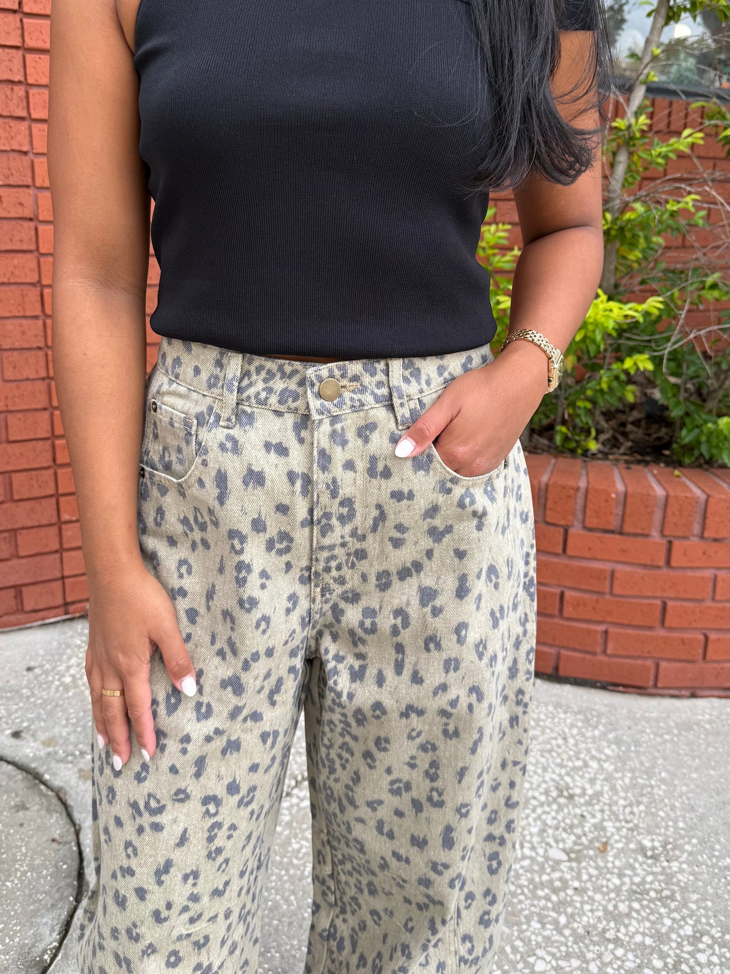 A person wearing beige wide-leg jeans with a gray leopard print pattern, paired with a black ribbed tank top, standing outdoors in front of a brick wall.