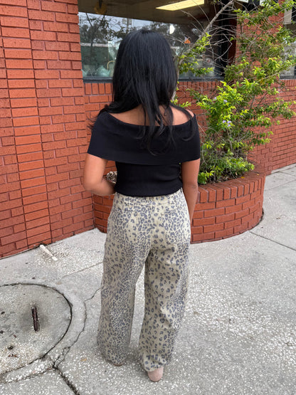 A woman viewed from behind wearing a black off-the-shoulder short-sleeve top and beige leopard-print wide-leg pants, standing on a sidewalk next to a red brick building.