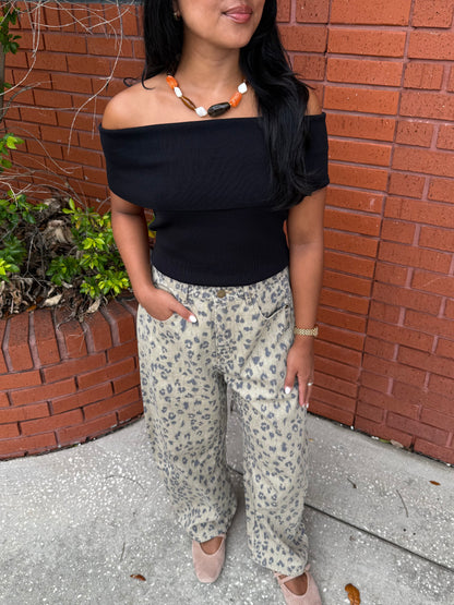 A woman wearing a black off-the-shoulder short-sleeve top and beige leopard-print wide-leg pants, standing outdoors against a red brick wall.