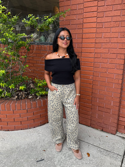 A woman stands outdoors against a red brick wall wearing a black off-the-shoulder top and beige wide-leg pants with a small dark floral print, paired with nude flats and sunglasses.