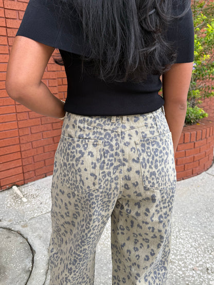 A person shown from the back wearing high-waisted beige pants with a gray leopard print pattern and a black ribbed short-sleeve top, standing outdoors against a red brick wall.