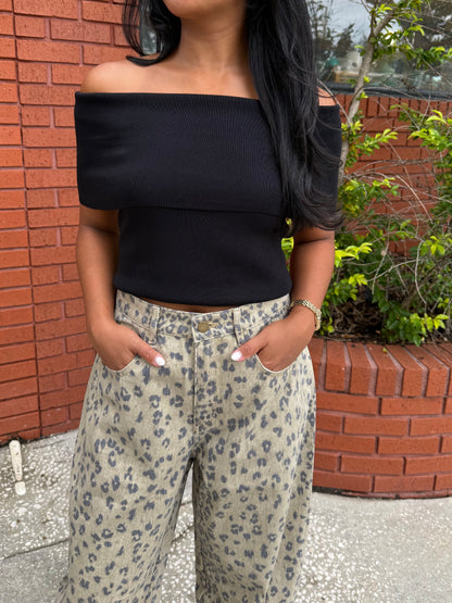 A woman wearing a black off-the-shoulder ribbed crop top and beige wide-leg pants with a gray leopard print, standing outdoors in front of a red brick wall.