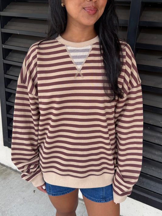 A woman wearing a brown and beige horizontally striped long-sleeve knit sweater with a V-neckline detail, paired with denim shorts, standing in front of a black slatted wall.