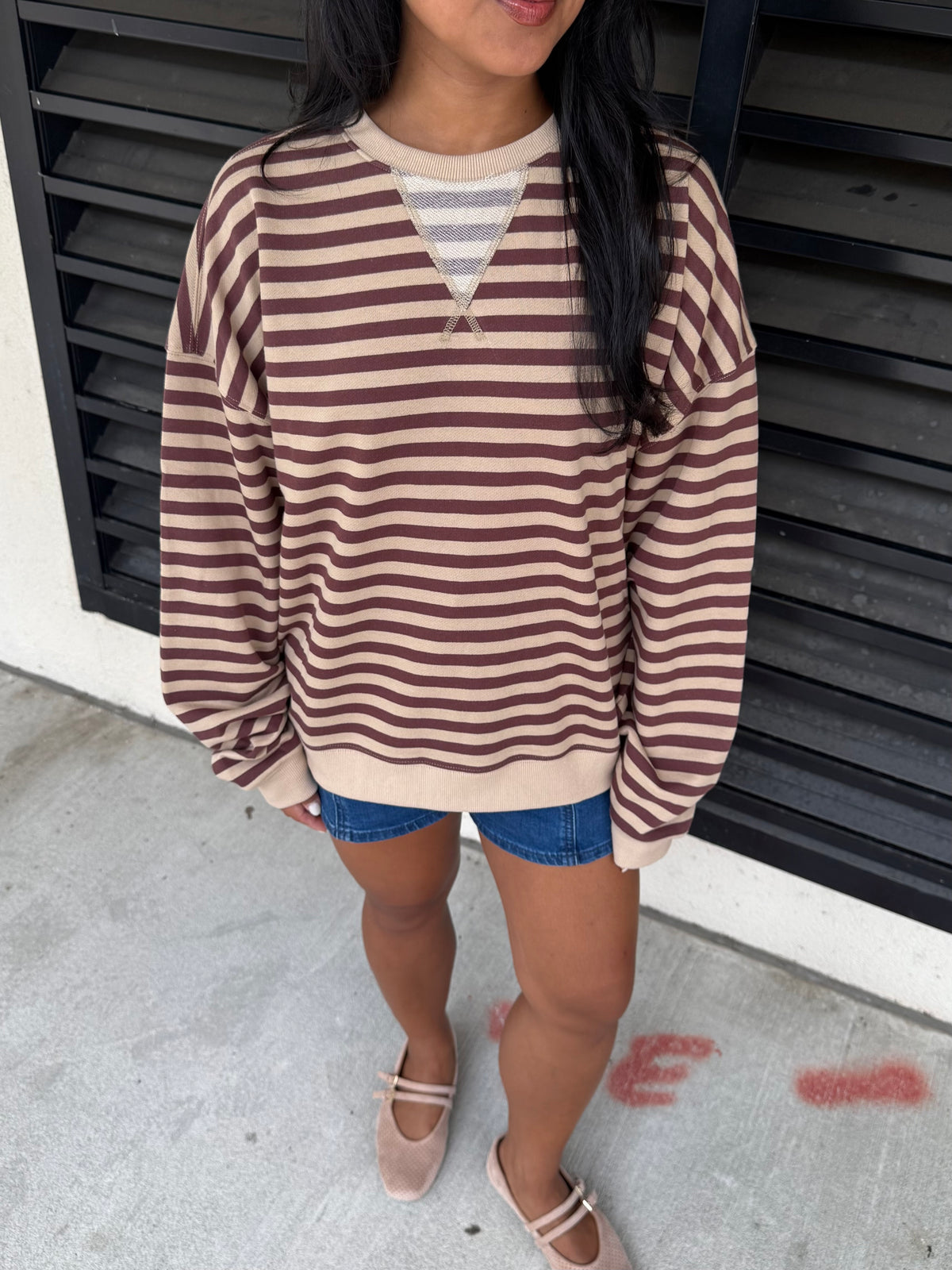 A woman wearing a brown and beige horizontally striped long-sleeve sweater over a gray striped V-neck undershirt, paired with denim shorts and beige Mary Jane shoes, standing in front of a black slatted wall.