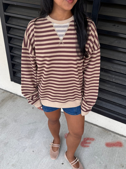 A woman wearing a brown and beige horizontally striped long-sleeve sweater over a gray striped V-neck undershirt, paired with denim shorts and beige Mary Jane shoes, standing in front of a black slatted wall.