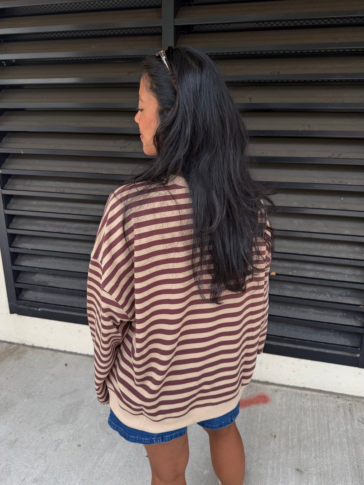 A woman with long black hair is shown from behind, wearing an oversized long-sleeve striped shirt with horizontal brown and beige stripes, paired with denim shorts, standing in front of a dark gray metal shutter.