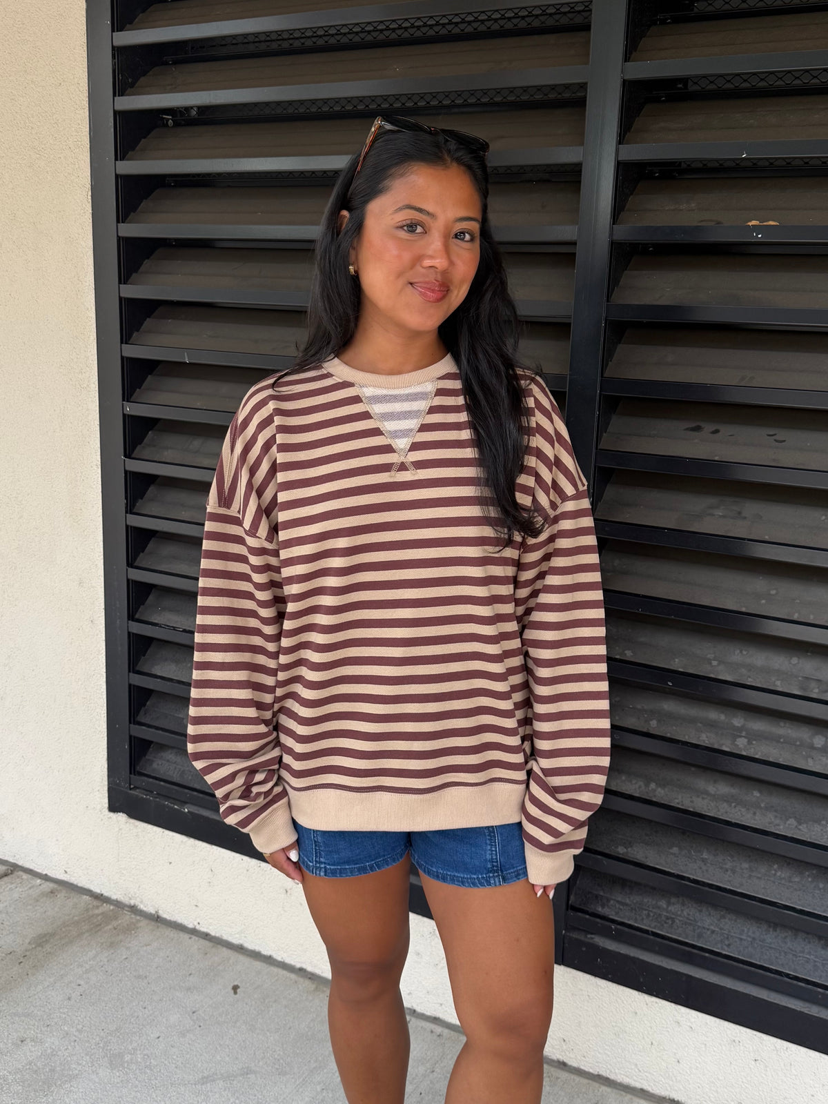 A woman wearing a brown and beige horizontally striped long-sleeve sweatshirt with a split V-neck over a white striped undershirt, paired with denim shorts, standing in front of a black metal shutter.