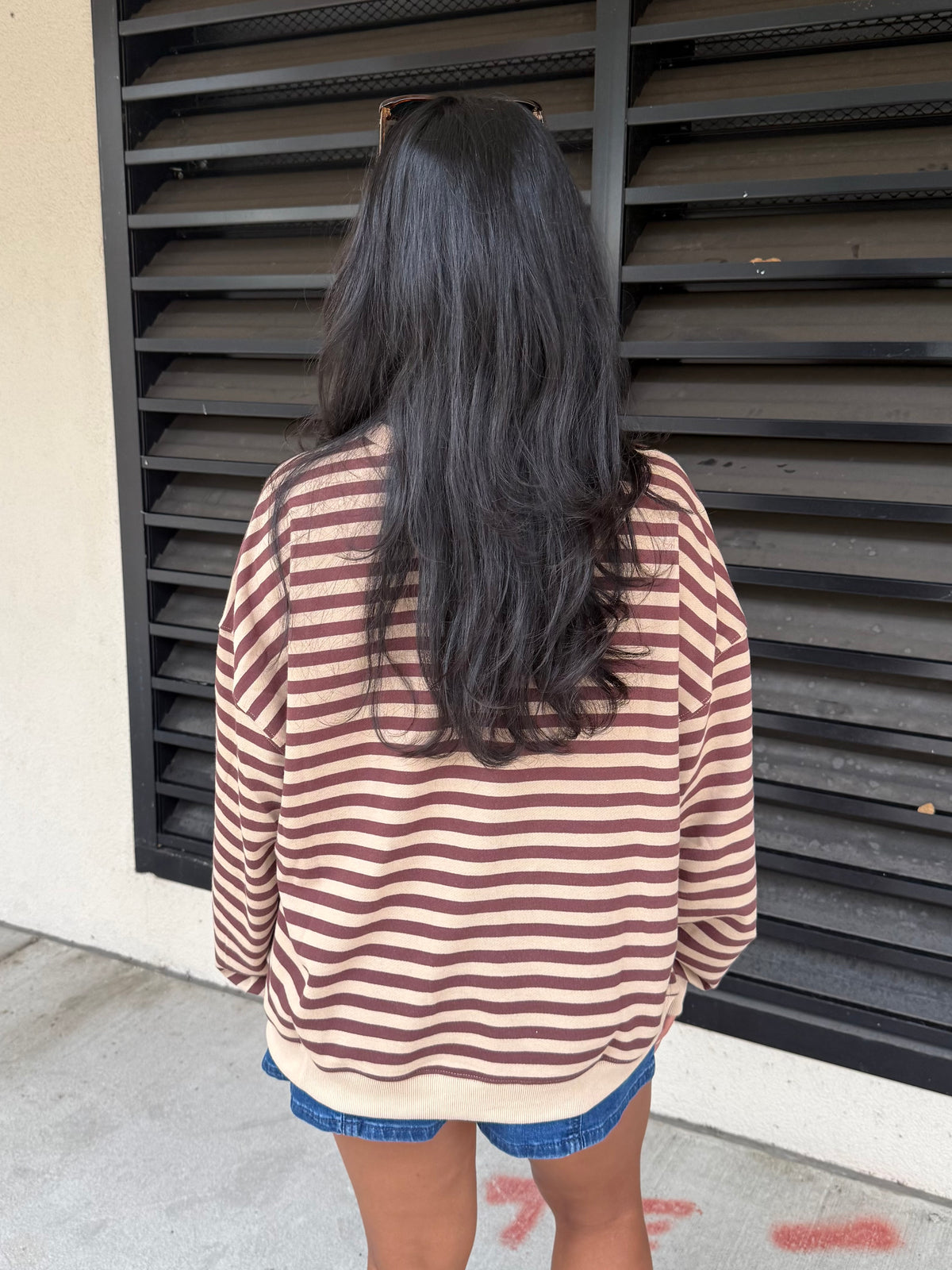 A person viewed from behind wearing an oversized long-sleeve top with horizontal rust-brown and cream stripes, paired with denim shorts, standing in front of a black metal shutter.