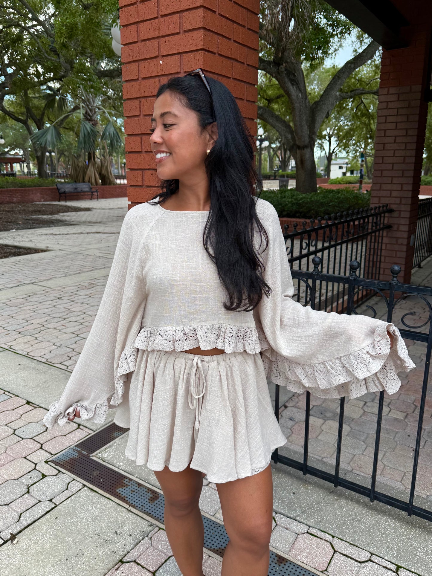 A woman wearing a two-piece cream-colored textured knit set with a long-sleeve crop top and matching ruffled shorts, standing outdoors in front of a brick column and black metal fence.