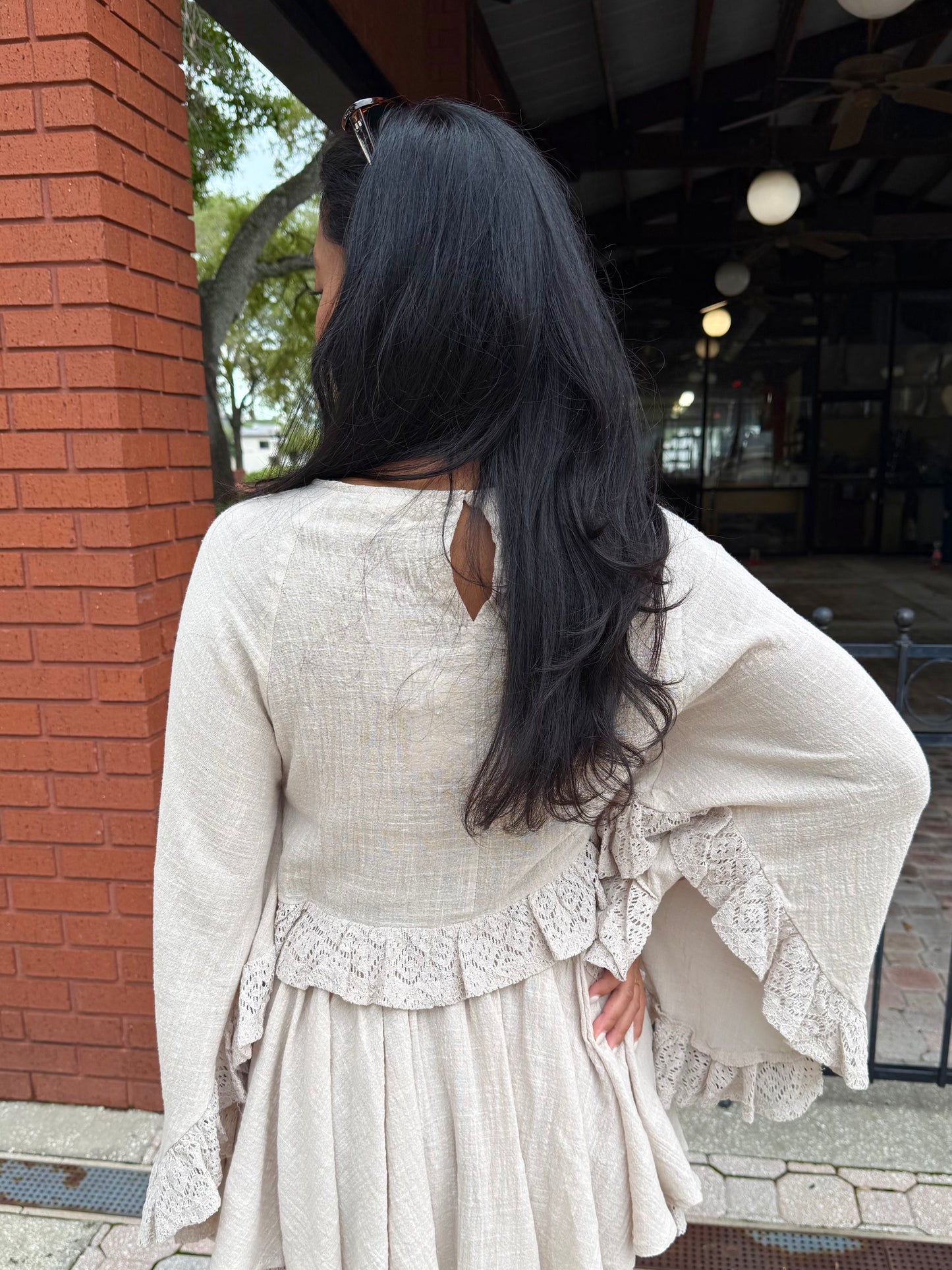 A woman viewed from behind wearing a cream-colored textured dress with lace ruffle details on the back and flared bell sleeves, standing outdoors next to a red brick column.