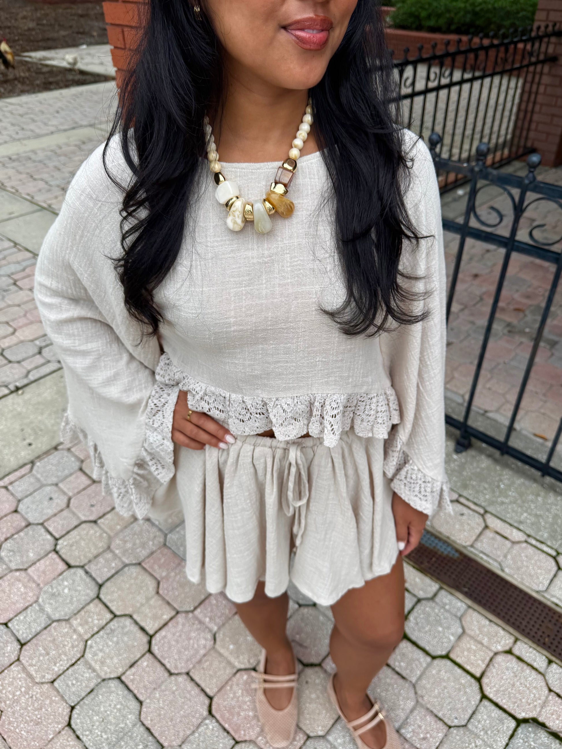 A woman wearing a cream-colored two-piece outfit consisting of a long-sleeve top with lace ruffle trim and matching shorts, styled with a chunky statement necklace and beige flat shoes, standing on a paved outdoor walkway.