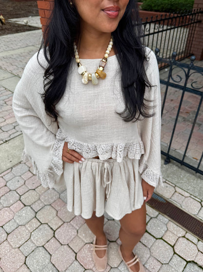 A woman wearing a cream-colored two-piece outfit consisting of a long-sleeve top with lace ruffle trim and matching shorts, styled with a chunky statement necklace and beige flat shoes, standing on a paved outdoor walkway.