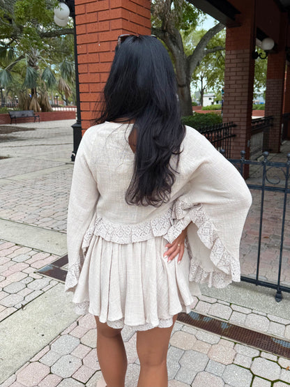 A woman viewed from behind wearing a cream-colored textured mini dress with long bell sleeves and lace ruffle trim, standing outdoors on a paved walkway near a brick building.