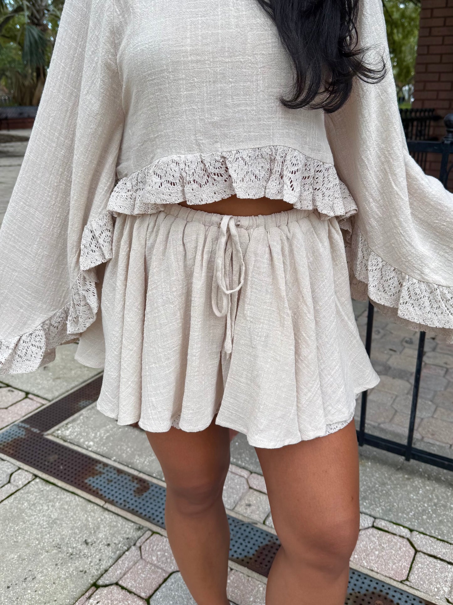 A person wearing a matching two-piece outfit with a cream-colored textured top and shorts featuring lace ruffle trim and a drawstring waist, photographed outdoors on a paved walkway.