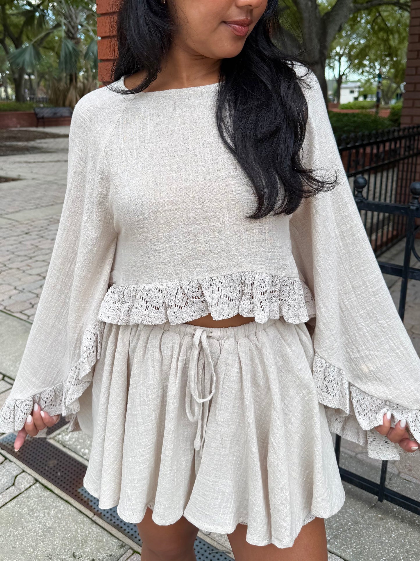 A woman wearing a light beige two-piece linen set featuring a long-sleeve crop top with lace ruffle trim and a matching drawstring mini skirt, photographed outdoors on a paved walkway.