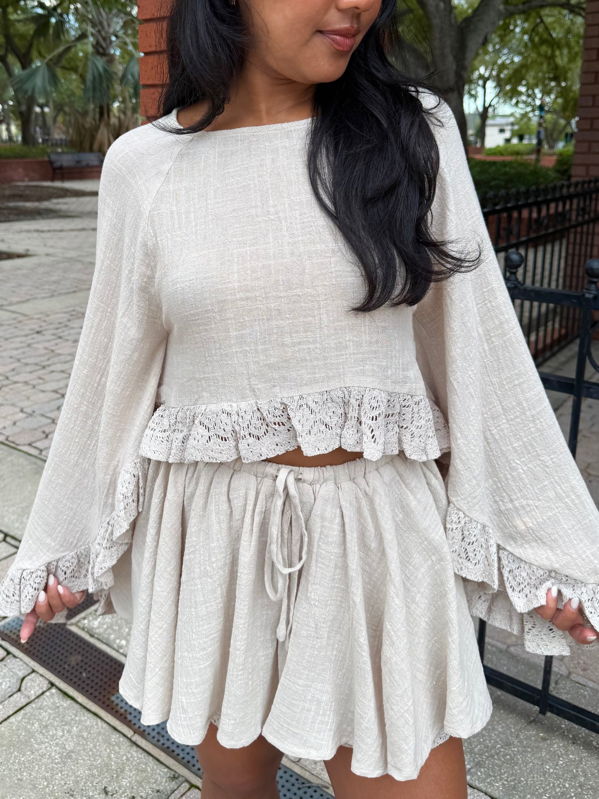 A woman wearing a light beige two-piece linen set featuring a long-sleeve crop top with lace ruffle trim and a matching drawstring mini skirt, photographed outdoors on a paved walkway.