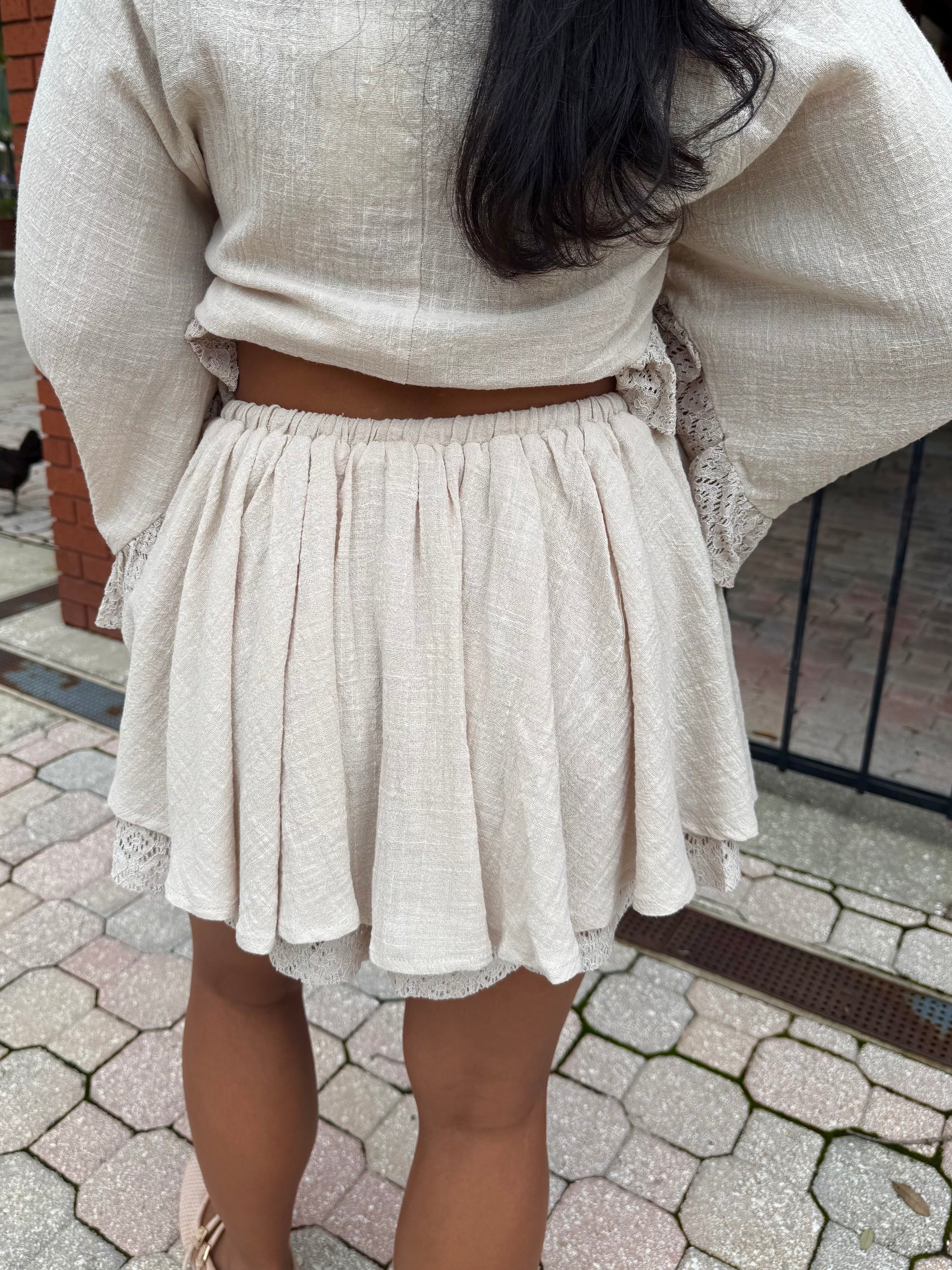A person shown from the back wearing a light beige textured two-piece outfit with a long-sleeve crop top and a matching pleated mini skirt, standing outdoors on a paved walkway.